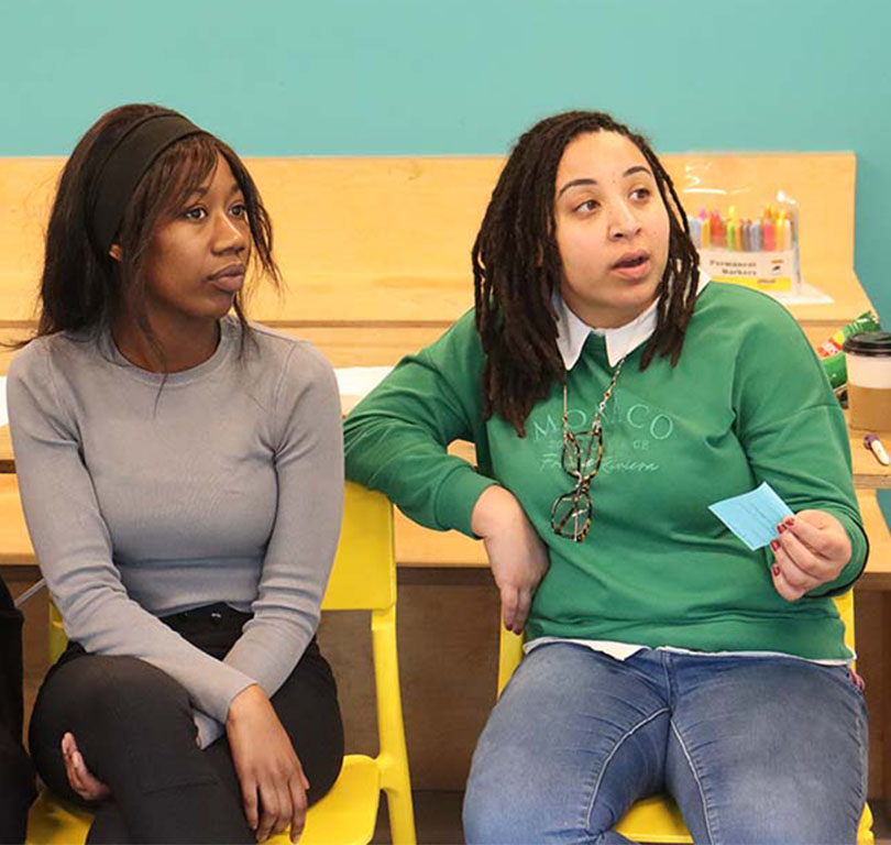 An image of two Black people sat down, with the person on the right speaking to someone to their left, out of view of the camera.
